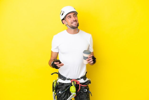 Junger Kletterer isoliert auf gelbem Hintergrund mit Kaffee zum Mitnehmen und einem Handy, während er etwas denkt