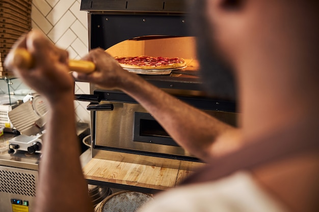 junger Koch, der vor dem Ofen steht und Pizza hineinlegt, während er im Restaurant arbeitet?