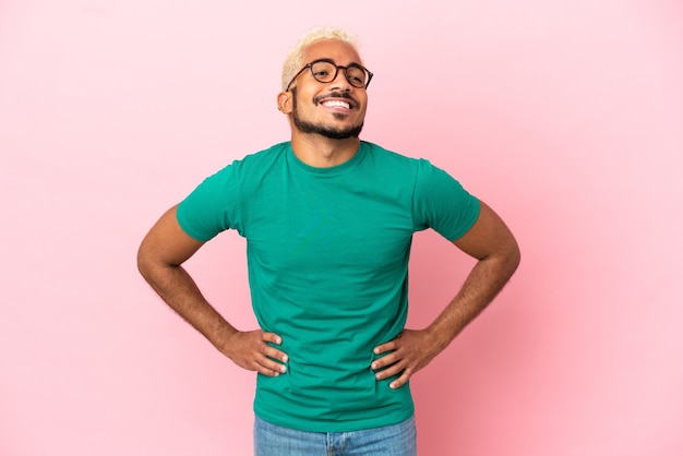 Junger kolumbianischer gutaussehender Mann isoliert auf rosa Hintergrund posiert mit Armen an der Hüfte und lächelt