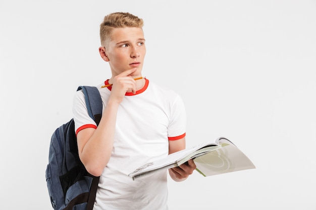 junger konzentrierter männlicher Student, der Rucksack trägt, der denkt und beiseite schaut, während er mit Lehrbüchern und Stift in den auf Weiß lokalisierten Händen studiert