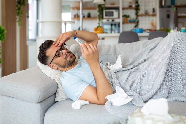 Junger kranker bärtiger Mann niest zu Hause auf grauem Sofa mit Decke in Serviette Krankheitsschutz Coronavirus-Viruskrankheit Grippe-Atemverband