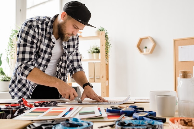 Junger kreativer Mann in der Freizeitkleidung, die sich über Arbeitsplatz beugt, während Werkstücke für dekorative Haustierhalsbänder im Studio vorbereitet werden