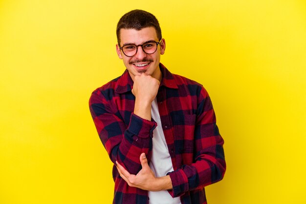 Junger kühler Mann lokalisiert auf gelber Wand, die glücklich und zuversichtlich lächelt und Kinn mit Hand berührt
