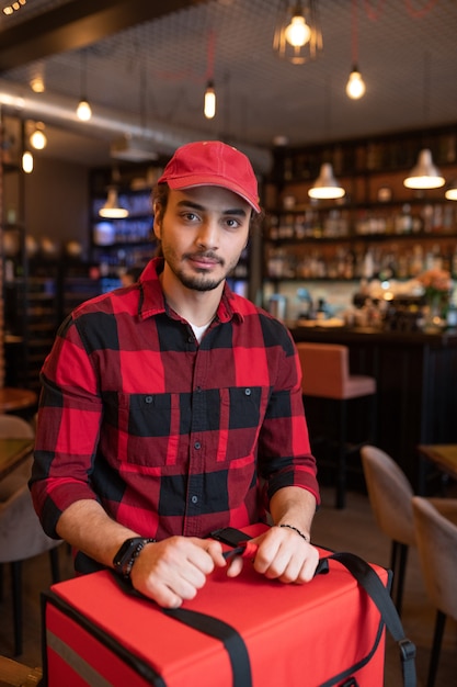 Junger Kurier des Café-Lieferservices, der durch große rote Tasche vor der Kamera steht, während er Bestellungen von Kunden liefert