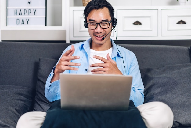 Junger lächelnder asiatischer Mann, der unter Verwendung des Laptop-Computer-Arbeits- und Videokonferenzmeetings Online-Chat zu Hause entspannt