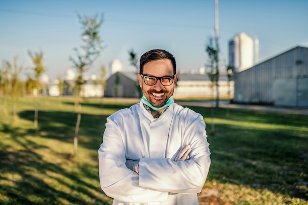 Junger lächelnder attraktiver kaukasischer Agronom in der Schutzuniform, die im Obstgarten steht