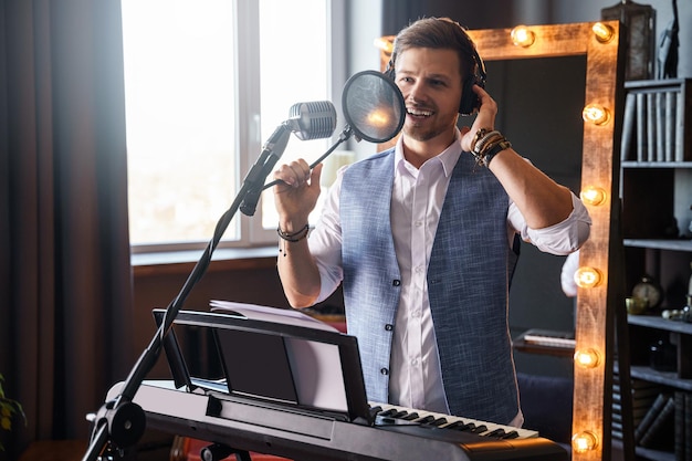 Junger lächelnder bärtiger Mann singt in einem Heimmusikstudio vor einem Klavier mit einem Schminkspiegel hinter seinem Rücken in ein Mikrofon