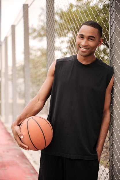 Junger lächelnder Basketballspieler, der im Park steht, während Basketball hält