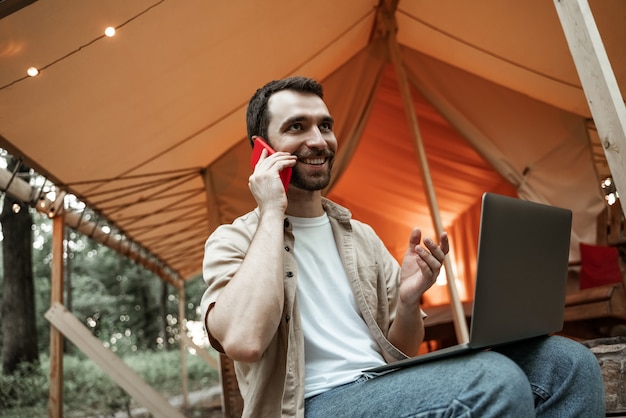 Junger lächelnder Brunettemann, der auf Glamping mit Laptop sitzt und auf Smartphone spricht und erklärt. Camping-Lebensstil. Reisen mit geringem Budget. Konzept der Remote-Arbeit. Moderne Internet-Kommunikationstechnologie.