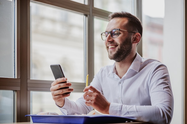 Junger lächelnder geeky Mann elegant gekleidet sitzend im Fast-Food-Restaurant, mit Smartphone und Pommes frites.