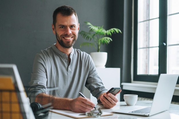 Junger lächelnder Geschäftsmann im lässigen grauen Longsleeve, der im dunklen, modernen Büroraum am Laptop arbeitet