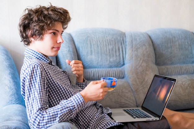 Junger lächelnder Jugendlicher mit dem langen Haar zu Hause studierend unter Verwendung des Laptops, der auf einem Sofa sitzt