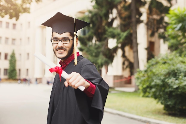 Junger lächelnder Mann an seinem Abschlusstag an der Universität mit Diplom. Bildungs-, Qualifikations- und Kleiderkonzept.