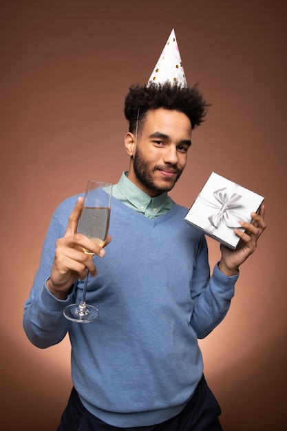 Foto junger lächelnder mann aus gemischten rennen in eleganter freizeitkleidung und geburtstagsmütze, der eine flöte mit prickelndem champagner und eine geschenkbox vor der kamera hält