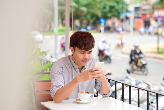 Junger lächelnder Mann, der am Telefon schaut oder eine Nachricht im Café im Freien liest