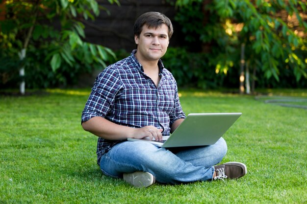 Junger lächelnder Mann, der auf Gras sitzt und mit Laptop arbeitet