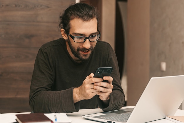 Junger lächelnder Mann, der eine Nachricht am Telefon schreibt, während er an seinem Arbeitsplatz sitzt