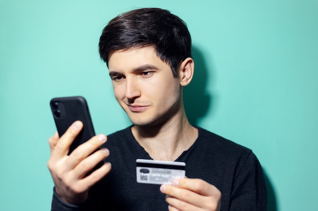 junger lächelnder Mann, der online durch Smartphone und Kreditkarte an der Wand von aqua menthe Farbe einkauft.