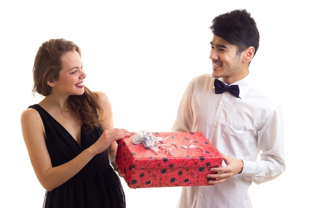Junger lächelnder Mann mit dem schwarzen Haar und junge hübsche Frau mit dem langen blonden Haar, das rotes Geschenk zeigt