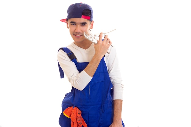 Junger lächelnder Mann mit dunklem Haar in weißem Hemd und blauem Overall mit orangefarbenen Handschuhen und blauem Snapback mit Schraubenschlüssel auf weißem Hintergrund im Studio