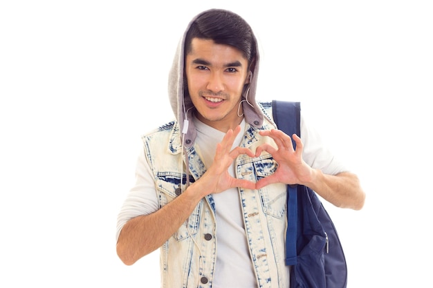 Junger lächelnder Mann mit schwarzen Haaren in weißem T-Shirt und Jeansjacke mit blauem Rucksack mit Kopfhörern und Herz auf weißem Hintergrund im Studio.