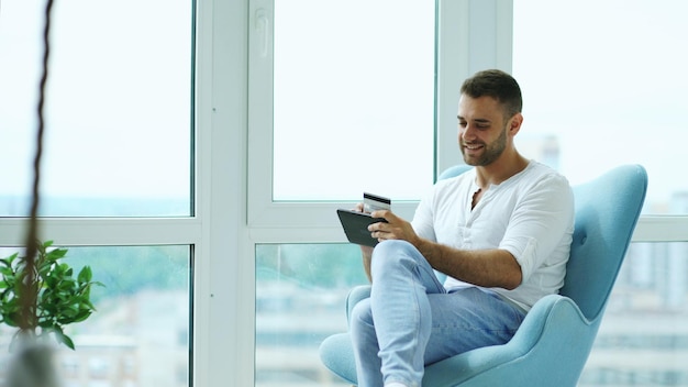 Junger lächelnder Mann Online-Shopping mit einem digitalen Tablet-Computer, der in einer modernen Loft-Wohnung sitzt