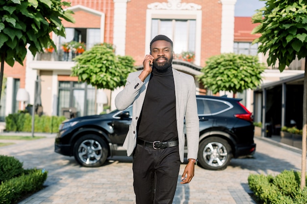 Junger lächelnder schwarzer Geschäftsmann in einem Anzug, der durch Smartphone spricht, das draußen nahe einem schwarzen Auto und modernen Backsteingebäuden geht