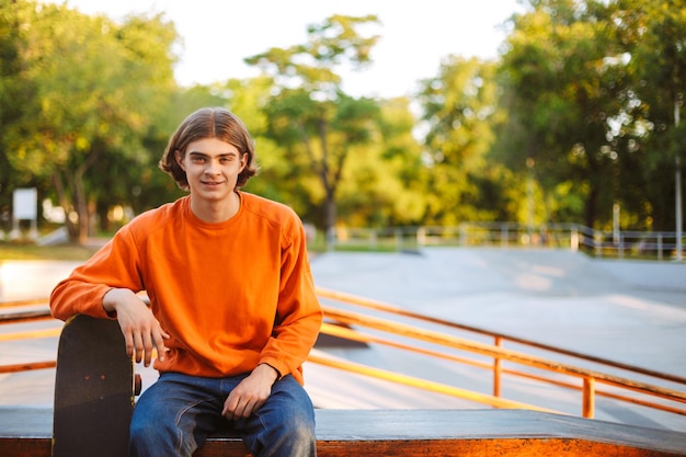 Junger lächelnder Skater im orangefarbenen Pullover, der freudig in die Kamera schaut, während er sich auf Skateboard mit modernem Skatepark im Hintergrund lehnt