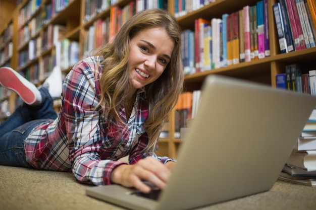 Junger lächelnder Student, der auf Bibliotheksboden unter Verwendung des Laptops liegt