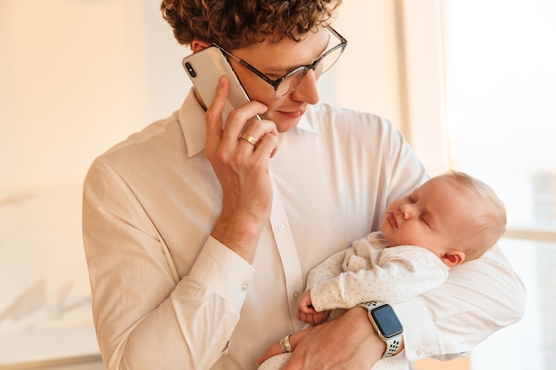 Junger lächelnder Vatergeschäftsmann, der mit seinem kleinen Babysohn spielt, während er zu Hause mit dem Handy spricht