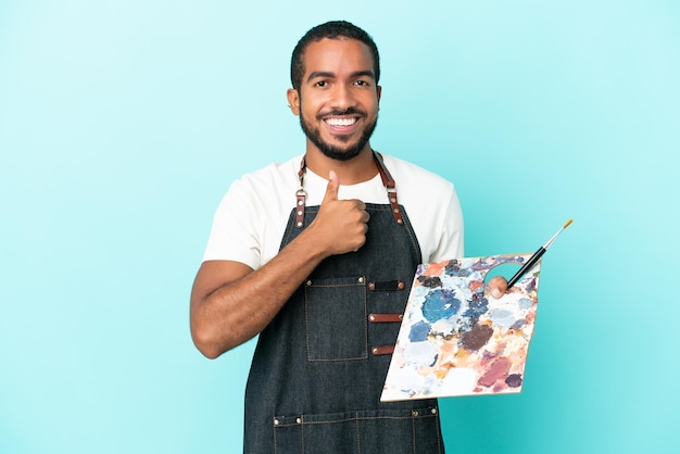 Junger lateinamerikanischer Künstler, der eine auf blauem Hintergrund isolierte Palette hält und eine Geste mit dem Daumen nach oben gibt