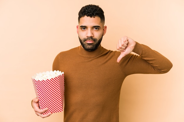 Junger lateinamerikanischer mann, der ein popcorn isoliert hält, das eine abneigungsgeste zeigt, daumen nach unten. uneinigkeit konzept.