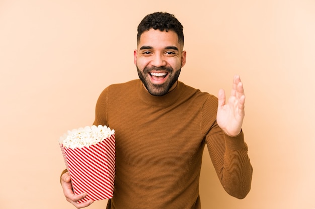 Junger lateinamerikanischer Mann, der ein Popcorn isoliert hält, das eine angenehme Überraschung empfängt, aufgeregt und Hände hebt.