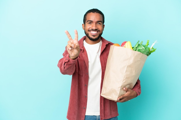 Junger lateinamerikanischer Mann, der eine Lebensmitteleinkaufstasche isoliert auf blauem Hintergrund hält, lächelt und Victory-Zeichen zeigt