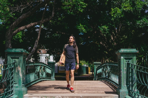 Junger lateinamerikanischer Mann mit langen Haaren und einer intelligenten Uhr, der eine Holzbrücke in einem Stadtpark mit einer Yogamatte auf dem Rücken passiert