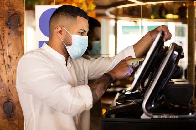 Junger lateinamerikanischer Restaurantangestellter mit medizinischer Maske, der die Rechnung am Kassenautomaten vorbereitet.