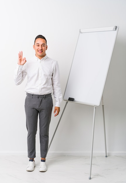 Junger lateinamerikanischer Trainer mit einem fröhlichen und selbstbewussten Whiteboard, das ok Geste zeigt.