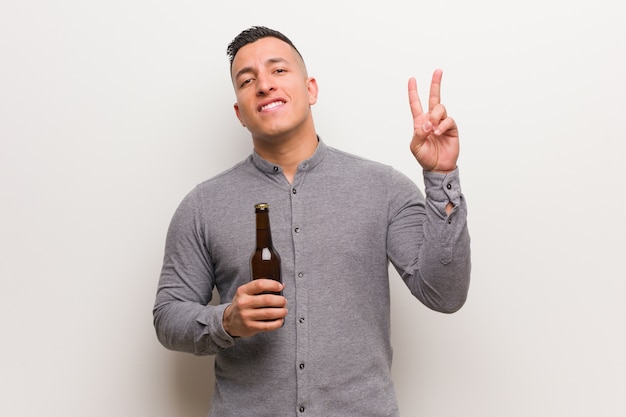 Junger lateinischer Mann, der ein Bier zeigt Nummer zwei hält
