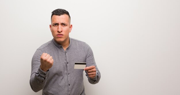 Junger lateinischer Mann, der eine Kreditkarte zeigt Faust zur Frontseite, verärgerter Ausdruck anhält