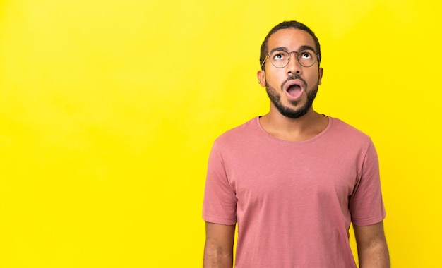 Junger lateinischer Mann isoliert auf gelbem Hintergrund, der nach oben schaut und mit überraschtem Ausdruck