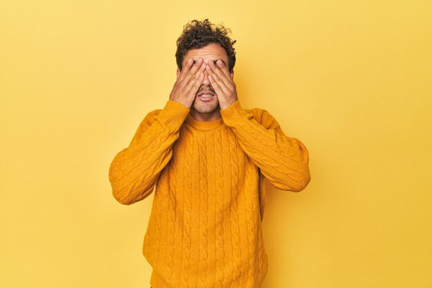 Junger Latino-Mann posiert auf gelbem Hintergrund und hat Angst, die Augen mit den Händen zu bedecken