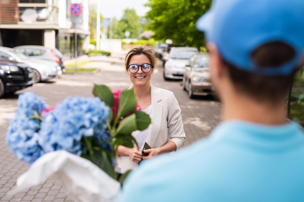 Junger Lieferbote in blauer Uniform liefert Blumen an eine Frau c