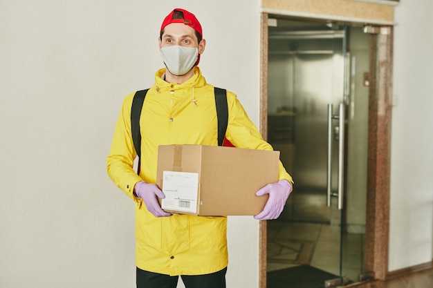 Junger Lieferbote in der gelben Jacke, die Maske und Handschuhe trägt und Paket im Bürokorridor hält