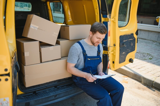 Junger Liefermann-Kurier in Uniform hält Dokumente in der Zwischenablage und überprüft die Paketpostfächer in der Nähe eines Autos für den Serviceversand an den Kunden Online-Shopping-Service-Konzepte