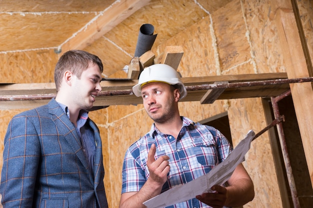Junger männlicher Architekt und Bauarbeiter Vorarbeiter Beratung von Bauplänen im unfertigen Haus mit freiliegenden Sperrholzplatten