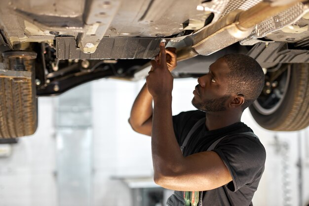 Junger männlicher Automechaniker in Uniform, die Auto im Autodienst mit angehobenem Fahrzeug prüft