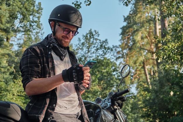 Junger männlicher Biker im Helm mit Handy und lächelnd auf einem Motorrad sitzend