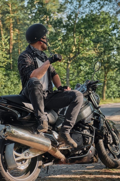 Junger männlicher Biker mit Helm fährt allein auf einem Motorrad, das an der Seite eines Waldes anhält und raucht