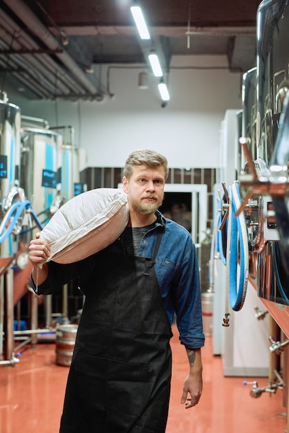 Junger männlicher Brauer in Arbeitskleidung, der einen weißen Sack voller Gerste auf der Schulter trägt, während er sich zwischen zwei Reihen riesiger Stahltanks mit Bier bewegt?