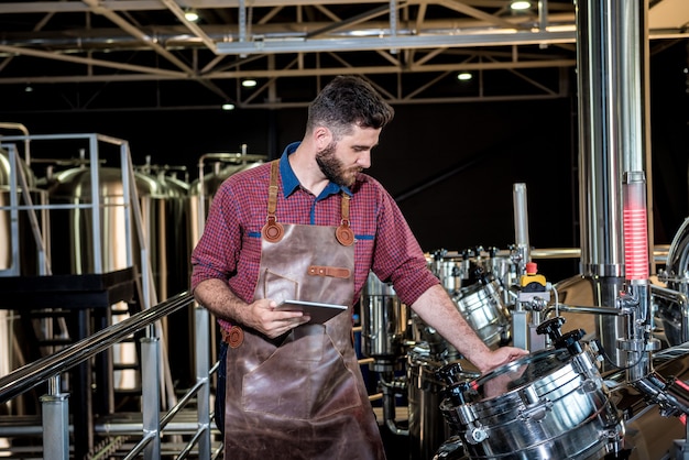 Junger männlicher Brauer in Lederschürze, der den Prozess der Biergärung in einem modernen Brauereibetrieb überwacht.